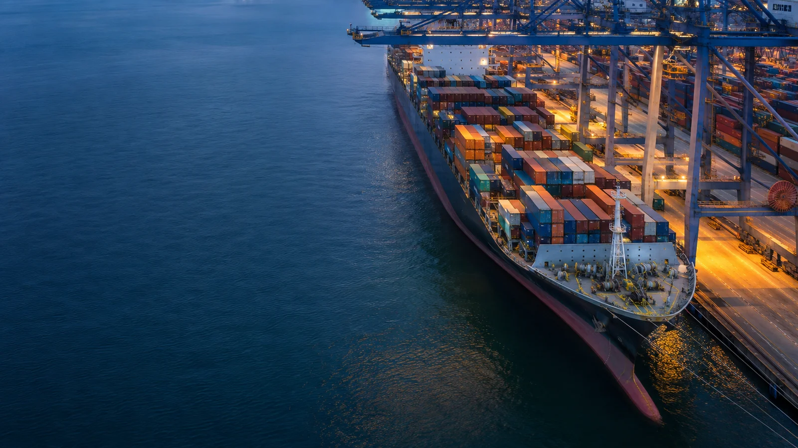 Container vessel beside tall port cranes and neatly stacked freight containers at dusk